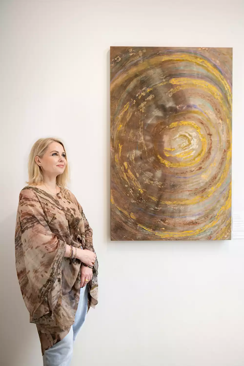 Artist Kasia Muzyka, stands beside her large, circular painting "Before First Breath," which has concentric rings of gold and brown