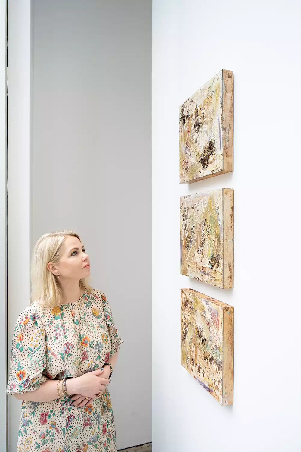 Artist Kasia Muzyka in a floral dress, standing in a gallery and looking at a series of her small, textured abstract paintings